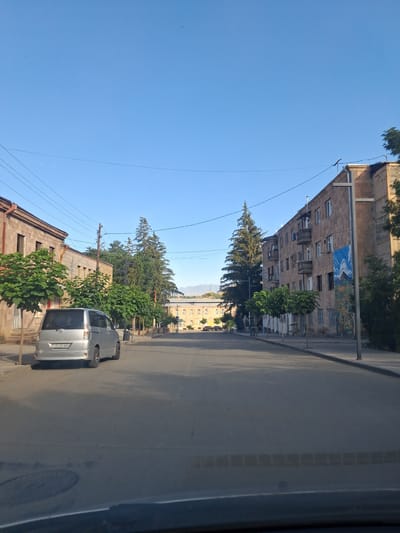 The image shows a street scene in the city of Vayk, Armenia, under a clear blue sky. The perspective is from inside a vehicle looking forward. On the left side of the street, there is a light brown, multi-story building with arched windows. In front of this building, several green trees with a rounded canopy are planted along the sidewalk. A silver minivan is parked on the right side of the street, facing away from the camera, with its license plate visible.

The street is paved with asphalt and appears to be in a residential or mixed-use area. On the right side of the street, there is a multi-story building constructed of light brown stone blocks. This building has several balconies with dark railings and numerous windows. A colorful mural depicting mountains and a blue sky is painted on the exterior wall of this building. More green trees are planted along the sidewalk in front of it.

In the distance, the street continues and appears to curve slightly to the left. Another building with a similar architectural style to the one on the left is visible in the distance, further down the street. Power lines are visible crisscrossing the sky, supported by utility poles. Tall, dark green coniferous trees are interspersed with deciduous trees along the street. In the far distance, mountains are faintly visible. The overall impression is a calm, daytime scene in a small city.