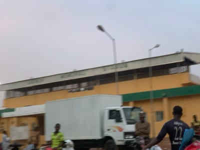 The image shows a white box truck parked in front of a yellow building with a green stripe near the bottom. The building has a large sign that reads "Katsina - _JUN" with part of the text obscured by the truck. There are people walking in front of the building, including one man wearing a green shirt and a young man wearing a blue shirt with the number "22" on the back. The scene is taken from a street-level perspective, with the building visible in the background. The coordinates provided locate the scene near the Ahmadu Bello Stadium in Sokoto, Nigeria. The sky is overcast and there are streetlights visible.