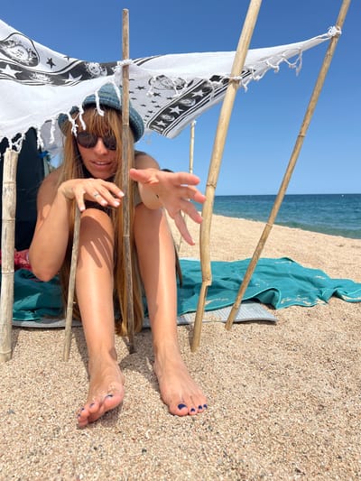 A woman sits on a sandy beach in Sant Pol de Mar, Spain, under a makeshift canopy constructed of bamboo poles and a patterned white fabric. She wears sunglasses and a denim hat, with her long blonde hair falling forward. Her right hand is extended towards the camera, palm open. Beside her, a teal towel is spread on the sand. The sea and a clear blue sky are visible in the background.