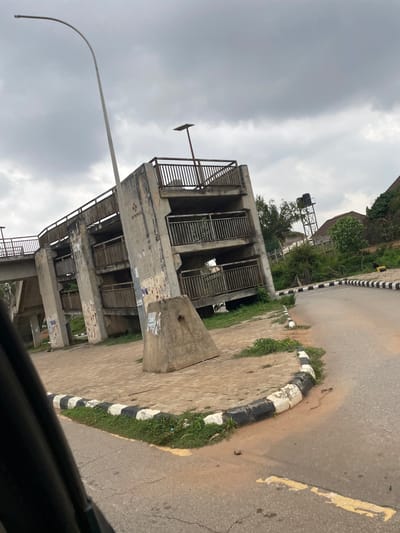 The image shows a concrete pedestrian bridge with multiple levels and ramps. The bridge is located in Ade, Nigeria, as indicated by the presence of a water tower in the background, which is characteristic of the region. The structure appears weathered and somewhat dilapidated, with visible signs of age and wear on the concrete and metal railings. The bridge is situated next to a paved road with a distinct black and white striped curb. Overcast, cloudy skies dominate the upper portion of the image, suggesting a potentially rainy or humid climate. Green vegetation, including grass and trees, is visible around the base of the bridge and along the roadside. The scene appears to be devoid of people. A street lamp stands tall on the left side of the frame, its curved pole extending towards the sky. The overall impression is one of urban infrastructure in a state of disuse or under heavy weather conditions.