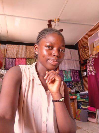 A young female subject is depicted in a medium shot, positioned indoors within what appears to be a textile or fabric shop located in the city of Ayob, Nigeria. She has dark skin, braided hair pulled back from her forehead, and small white stud earrings. She is wearing a light peach sleeveless collared top with subtle embellishments on the shoulders and a multi-bead bracelet on her left wrist. Her right hand is raised, with fingers gently touching her chin. She maintains direct eye contact with the camera, exhibiting a subtle smile and visible dimples on her cheeks.

The background on the left side of the frame features multiple rows of folded fabrics or textiles displayed on shelves, showcasing various patterns and colors including gold, brown, purple, pink, white, and green. On the right, a vertical wooden slatted wall panel is visible, with a gold circular decorative emblem or sign mounted upon it. Below this, a partial view of a mannequin torso, dressed in a dark red garment with distinctive white swirling patterns, is present. Additional folded fabrics are stacked on shelves beneath the mannequin. The ceiling overhead is white and paneled, featuring a light fixture with an exposed bulb and a brown ceiling fan motor.