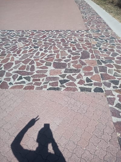 The image depicts an outdoor paved area in La Venta, Mexico. The ground surface is segmented into three distinct zones: a smooth, reddish-brown concrete slab in the upper left quadrant; a lower section comprised of reddish-brown interlocking pavers featuring a textured, undulating pattern; and a mosaic border composed of irregularly shaped, multicolored stones (red, brown, grey, black) set in light-colored mortar. This mosaic pattern separates the smooth concrete from the interlocking pavers and extends along the right edge of the frame. A narrow, light-colored concrete strip and a patch of dry, light green grass are visible in the upper-right background. A dark, distinct human shadow is cast onto the lower-left portion of the interlocking pavers. The shadow's left arm is raised, with the hand forming a "V" gesture. The head-area silhouette includes a rectangular object held by the individual, consistent with a camera or smartphone being used to capture the image.