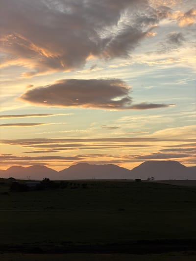 The image shows a landscape at sunset in what appears to be a rural area in or around George, South Africa. The sky is filled with layers of clouds, some dense and puffy, colored with orange and pink hues from the setting sun. Other clouds are thin and linear, stretched across the sky. In the distance, there are mountain ranges visible, with their silhouettes against the sky. The foreground consists of a dark green field with a building in the lower center with power lines on the side of the mountain. It is not possible to see any people in the image.