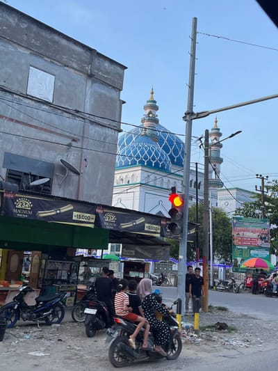 The image depicts a street scene in Dayahlhoksukon Barat, Indonesia, with a prominent mosque in the background. In the foreground, several people are present, with some individuals riding motorcycles. The environment appears to be a bustling street with shops and advertisements visible.
Specifically, the mosque features distinctive blue domes with white ornate patterns, and two tall minarets. In front of the mosque, a traffic light shows a red signal. To the left of the traffic light, a building with a gray facade has a white, possibly reflective, covering over a window. Banners displaying advertisements for "MAGNUM KRETEK" are hung across the front of a shop. Further to the right, a larger banner announces "PANITIA QURBAN Menerima & Menyalurkan HEWAN QURBAN," indicating a collection for animal sacrifice, with prices listed for individuals.
Several motorcycles are parked or in motion on the street. People are seen in various poses: some standing, others seated on motorcycles. The ground is paved with asphalt and appears somewhat littered. Trees and utility poles are also part of the urban landscape. The sky is a clear blue, suggesting daytime.