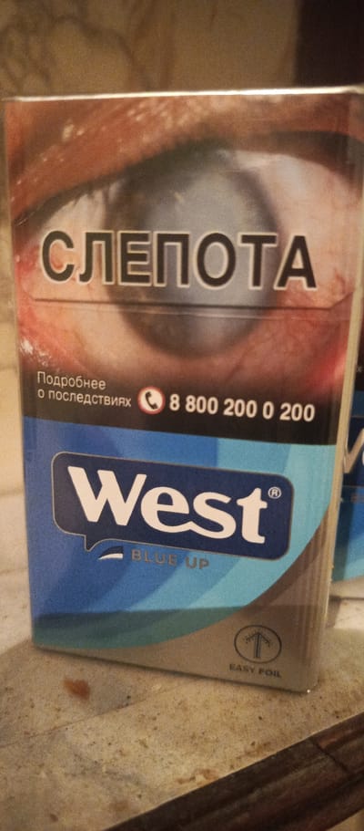 This is a close-up, slightly angled photograph of the front of a cigarette pack. The pack features a stark image of an eye with what appears to be a cataract, conveying a warning. In large, bold, black letters, the Russian word "СЛЕПОТА" (meaning "blindness") is prominently displayed across the top portion of the pack, overlaying the eye.

Below this warning, smaller text in Russian reads "Подробнее о последствиях" (meaning "More about the consequences"). To the right of this text, a phone icon is visible next to the number "8 800 200 0 200".

The lower half of the pack is dominated by a blue color scheme. A prominent white logo, "West", is presented in a speech bubble-like graphic. Beneath the "West" logo, the words "BLUE UP" are printed in a lighter blue. In the bottom right corner of the pack, a small graphic of an arrow pointing upwards within a circle is visible, with the text "EASY FOIL" printed below it.

The lighting in the image appears to be artificial, casting a warm tone on the surrounding surface, which seems to be a textured, possibly stone or wood, countertop. The background is out of focus but suggests an indoor setting. The overall mood conveyed by the pack design is serious and cautionary, due to the prominent warning about blindness. The context of Polyarnye Zori, Russia, indicates the geographical origin of the product.