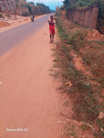 The scene depicts a roadside in the city of Umudioka, Nigeria. A young individual, dressed in a red short-sleeved top with white accents and red shorts, stands facing the camera on a reddish-brown dirt path. Their arms are crossed over their chest. To the left of this path, a paved, dark grey asphalt road extends into the background. In the mid-ground on this road, a motorcyclist is visible, moving away from the camera.
Along the right side of the dirt path, a strip of unkempt green vegetation, dry brush, and scattered refuse is present. Behind this vegetation, a low, weathered stone or concrete wall, partially covered by foliage, runs parallel to the road. The left side of the road in the background shows signs of partial construction or development and dense green foliage. The sky is a pale blue-grey. The watermark "Galaxy A52s 5G" is visible in the lower-left corner of the image.
