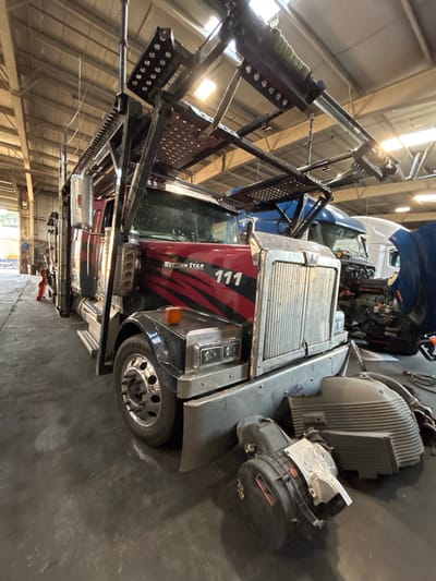 This media file shows the interior of a commercial vehicle repair shop in Aurora, United States of America. The main subject is a large truck, likely a semi-truck, with its hood open. The truck has a red and black decal on its side with the number "111" prominently displayed. The truck is also branded with "WESTERN STAR". The scene is indoors, as indicated by the metal ceiling structures and fluorescent lighting. Several components of the truck's engine are visible, as well as other parts and equipment typical of a repair facility, such as hydraulic lifts and metal racks. The floor is concrete. The image appears to be taken during daylight hours, suggested by the bright natural light filtering in from what seems to be a large garage door or windows. No people are visible in the frame, and the overall atmosphere is one of maintenance and repair work. The image is oriented vertically.