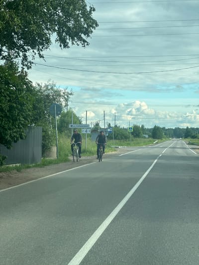 Two people on bicycles are riding on the side of a paved road. The road has a white line marking the lane. The scene is outdoors with a cloudy sky. Trees and greenery are visible on the left side of the road, and a line of trees stretches into the distance on the right. There are several signs visible on the left side of the road, including a blue sign with white text that reads "ул. КАМЫШОВАЯ" and a green rectangular sign with the numbers "2 4 6". There is also a pedestrian crossing sign. Power lines are visible in the sky, running parallel to the road. The overall impression is that of a rural or suburban setting. The time of day appears to be daytime, and the weather is overcast.