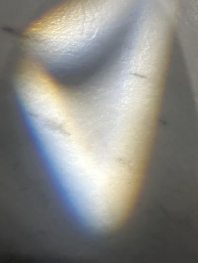 This media file is an abstract image that appears to capture the effect of light refracting through a prism or water. The primary subjects are bands of color, resembling a spectrum, that stretch across a textured, uneven surface. There are no visible people, recognizable objects, or clear scene or setting details.

The activity taking place is the interaction of light with a surface, creating a prismatic effect. Notable details include the presence of colors that suggest a rainbow or a similar optical phenomenon. The colors present are yellows, oranges, blues, and purples, blending into each other. The background surface is predominantly white or light grey, with subtle textures and what appear to be small dark specks, possibly dust or imperfections on the surface. The lighting conditions are not clearly discernible beyond the fact that light is present and interacting with the surface in a way that causes refraction. There are no location cues or emotional undertones that can be identified from the image itself. No text is visible in the media file.