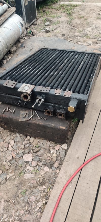 The image shows a close-up, high-angle view of a black radiator with a gridded surface, likely for an industrial or automotive application, lying on a wooden surface and next to a pile of gravel. Several metal plates and bolts are scattered around the radiator. To the left of the radiator, partially visible, is a large, cylindrical metallic object with blue text. In the background, a black case and some vegetation are visible. A red hose is visible on the wooden surface in the lower right corner. The setting appears to be an outdoor area, possibly a workshop or storage yard. The image does not contain any people or identifiable landmarks that would allow for a specific location within Rizunenkove, Ukraine.