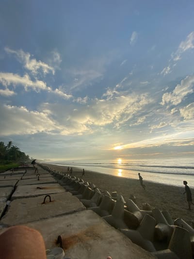 This image depicts a beach scene at sunrise in Bateetimoh, Indonesia. The foreground is dominated by a concrete sea barrier composed of interlocking tetrapods, some of which have rusted metal loops. A person's knee and foot are visible in the lower left corner of the frame, suggesting the photo was taken from a seated position.
Beyond the sea barrier, a sandy beach stretches along the coastline. Several people are present on the beach, some of whom appear to be playing. Further out, the ocean is calm with gentle waves reaching the shore. The sun is rising over the horizon, casting a golden reflection on the water and illuminating the sky. The sky itself is a blend of blues and whites, with scattered clouds catching the sunlight.
To the left of the beach, a line of palm trees and vegetation forms the edge of the land. In the distance, the coastline curves, disappearing into a hazy horizon. The overall atmosphere is serene and tranquil, capturing the early morning beauty of a tropical beach.