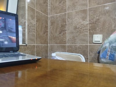 The image captures an indoor scene with a focus on a laptop situated on a wooden table. The laptop's screen is partially visible, displaying what appears to be a game or application. Two small orange indicator lights are illuminated on the laptop's casing. To the left of the laptop, a white spray can with a blue cap stands upright. Behind the laptop and spray can, a tiled wall with a brown, mottled pattern is visible. Further to the right, a light-colored chair with a blue trim is partially seen, with a clear plastic bottle with a white cap resting on it. Adjacent to the chair, a light switch is mounted on the tiled wall. On the far right of the image, a translucent plastic bag filled with items, including a blue and white object, is placed. The scene is likely within a residential or office space in Sabunchu, Azerbaijan, suggesting a personal or work environment.