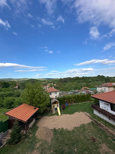 The image depicts a scene in Pchelin, Bulgaria. The view is from an elevated position, overlooking a residential area. The sky is blue with scattered white clouds. Houses with red tile roofs are visible, interspersed among trees and greenery. A playground with a swing set and a small playhouse is located in one of the yards. A section of the yard appears to have been recently filled with sand.