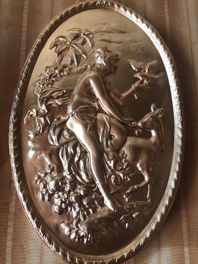 This is a close-up, top-down, indoor shot of an oval-shaped, metallic relief on a patterned cloth background. The relief depicts a mythological scene featuring a female figure, a deer, and flora. The woman, with her back to the viewer, is seated and appears to be interacting with the deer, possibly offering it a sprig of leaves. She is adorned with drapery and has her hair styled elaborately. The deer is depicted in profile, reaching towards the woman. Lush foliage, including what appear to be palm leaves and fruits, surrounds the figures. The metallic surface of the relief exhibits a high polish, reflecting light and creating highlights and shadows that accentuate the detailed craftsmanship. The cloth background features a repeating pattern of alternating beige and cream vertical stripes. The image does not provide any information or visual cues that would allow for the identification of a specific location in the city of Chiatura, Georgia.