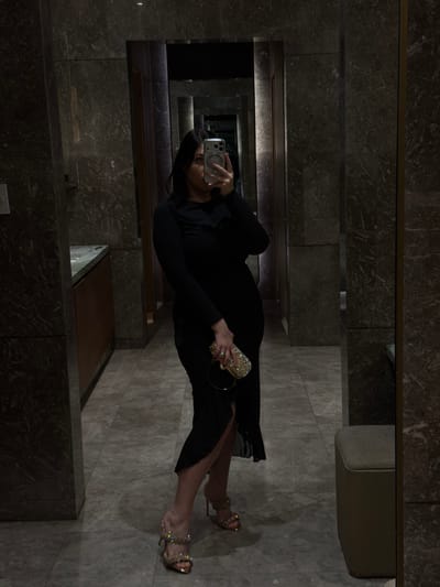 A person in a black dress stands in front of a mirror, taking a selfie. The person is wearing high heels and holding a jeweled clutch. The background appears to be a hallway with marble walls and floors, possibly in a hotel or a high-end establishment. There are reflections in the mirror showing more of the hallway. The lighting is dim and moody. There is no indication that this location is in Cebu City, Philippines.