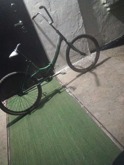 A low-angle, full shot shows a green bicycle leaning against a dark, textured wall. The bicycle has a white seat and is positioned diagonally across the frame. Its front wheel is positioned towards the upper right of the image, while the rear wheel is closer to the bottom left. The bicycle's frame is thin and curved, and the handlebars are narrow and upright. The ground beneath the bicycle is a mix of a textured, grey concrete surface and a green, carpet-like material. Shadows are cast on the ground by the bicycle and the wall, indicating a light source from the upper left. The overall scene is dimly lit, suggesting it may be evening or nighttime. The location is Leontyevskoye, Russia.