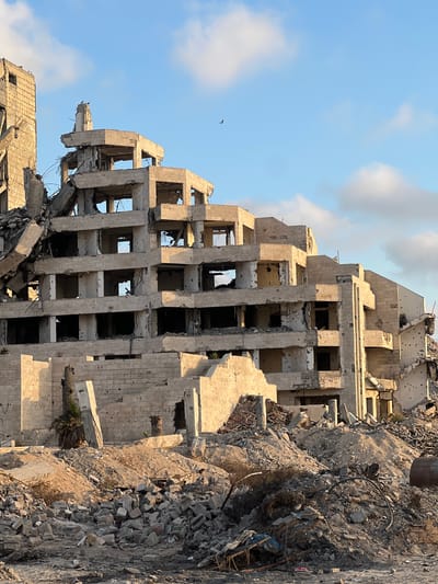 In Jabalia, Palestinian Territories, the skeletal remains of a multi-story concrete building stand amidst a landscape of rubble. The structure, once likely a hotel or apartment complex, exhibits severe damage, with gaping holes where windows and walls once were. Sections of the building have collapsed, exposing twisted rebar and shattered concrete. The facade is pockmarked with numerous small holes, suggestive of sustained shelling or gunfire. Debris, including shattered bricks, concrete fragments, and twisted metal, is piled high around the base of the building and across the foreground, obscuring any indication of its original surroundings. A single, sparse palm tree struggles to survive near the foundation, a small patch of green against the prevailing destruction. The sky above is a pale blue, dotted with a few wispy clouds, and a solitary bird can be seen in flight. The scene conveys a powerful sense of devastation and aftermath.
