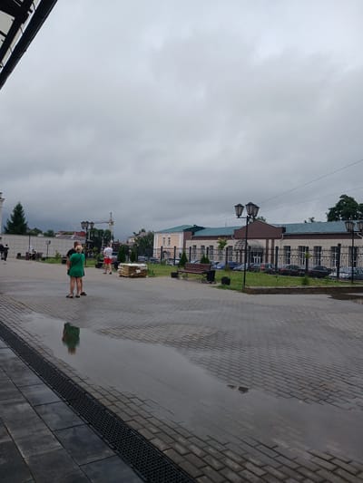 The image depicts a street scene in Michurinsk, Russia, under a cloudy sky. In the foreground, a wet cobblestone pavement is visible, with puddles reflecting the overcast conditions and the surrounding environment. A dark metal grate runs along the edge of the pavement.
Several people are present in the scene. In the middle ground, a woman in a green dress and shorts stands facing away from the camera, with her reflection visible in a puddle. Next to her, a man in a white t-shirt and red shorts is also standing. Further in the background, other individuals can be seen walking and sitting on benches.
Various objects and structures are scattered throughout the image. Lampposts with multiple lights are positioned along the street. A building with a beige facade and a dark roof is visible on the right side of the frame, with a black metal fence in front of it. Cars are parked behind the fence. In the distance, a construction crane is visible. Piles of what appear to be building materials are stacked on the pavement. Trees and greenery are interspersed throughout the scene. The overall atmosphere suggests a damp, possibly post-rain, day in an urban setting.
