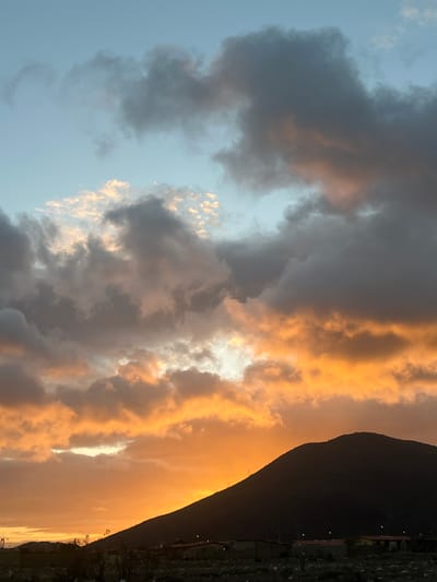 This image captures a dramatic sunset over a mountainous landscape, likely in the vicinity of San Antonio, Venezuela. The sky is filled with a vibrant display of clouds, ranging from dark, ominous grays to brilliant oranges and yellows, illuminated by the setting sun. The light of the sunset casts a warm glow, highlighting the textures and contours of the clouds.

In the foreground, a silhouetted mountain dominates the right side of the frame, its dark form contrasting sharply with the bright sky. At the base of the mountain, a semi-urban or rural area is visible. Scattered structures with red roofs and some dimly lit windows suggest human habitation. Some utility poles or towers are also present. The landscape below appears somewhat arid and undeveloped, with low-lying vegetation and rough terrain.

The overall impression is one of a rugged natural environment experiencing a spectacular atmospheric event. The image is a static, landscape shot taken from a ground-level perspective, emphasizing the scale of the mountain and the grandeur of the sky. No people or specific activities are depicted.