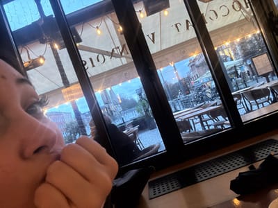 A low-angle indoor shot shows a person with long eyelashes, their face partially visible in the bottom left corner, fingers near their mouth. Outside, a streetscape is seen through multi-paned glass windows. The exterior view features tables and chairs in front of buildings, with umbrellas and bicycles visible. A sign reads "CHOCOLAT V." String lights are strung above the outdoor area. The overall tone is somewhat muted, with an overcast sky.