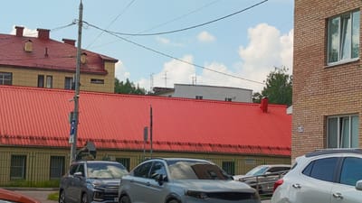 The image shows a street scene in Zvenigorod, Russia, dominated by a large building with a vibrant red corrugated metal roof. To the left of the red-roofed building, a lighter colored building with a red tiled roof and dormer windows is visible. In the foreground, several cars are parked or driving, including a silver SUV and a white SUV. A light pole with a blue parking sign is positioned near the center of the image. The sky is partly cloudy, with a clear blue expanse and fluffy white clouds. Trees are visible in the background, interspersed with other buildings. The overall impression is that of an urban or suburban environment on a sunny day.
