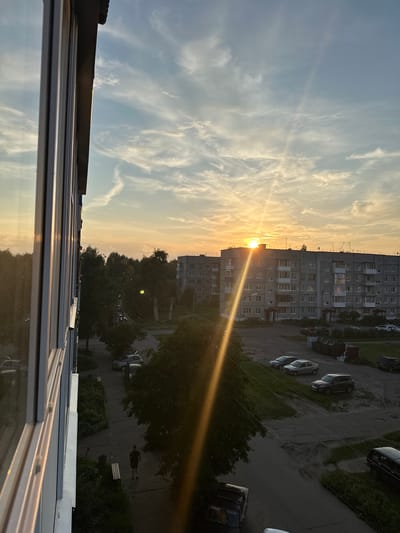 The image depicts a sunset over a residential area in Kotlas, Russia. The perspective is from a window of a multi-story building, looking out towards apartment blocks and trees. The sun is setting on the horizon, casting a warm, golden light across the sky and illuminating the clouds. A bright sunbeam streaks diagonally across the scene, from the sun towards the foreground.
In the foreground, the edge of a building with multiple windows is visible on the left side of the frame. The windows reflect the sky and the sunset. Below the building, a paved walkway runs alongside a strip of greenery and trees. A single person is walking on the walkway, silhouetted against the darkening environment.
Further back, more apartment buildings, characteristic of Soviet-era architecture, are visible. Cars are parked in a lot or on the street in front of the buildings. The overall atmosphere is one of a quiet evening in a typical urban residential district.