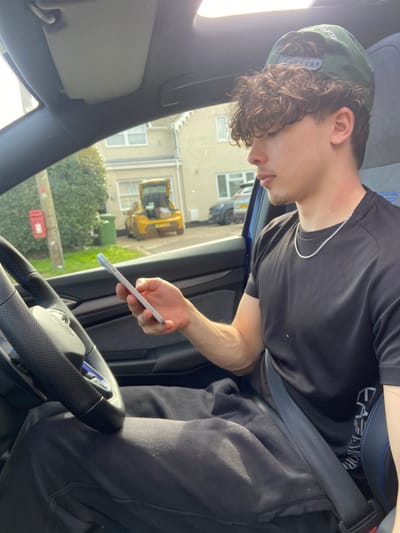 A young male is seated in the driver's seat of a vehicle on a residential street in Broxbourne, United Kingdom. He wears a dark green baseball cap reversed, a black t-shirt, a silver chain necklace, and black sweatpants. His right hand holds a light-colored smartphone, at which he is gazing downwards. He is secured by a black seatbelt. The vehicle's interior features a black, textured steering wheel with blue accents and blue trim on the seat. In the background, visible through the windshield, a light-colored residential house stands opposite. A yellow compact car with its rear hatch open, revealing contents, is parked in front of the house. Further down the street, a dark-colored SUV is also parked. A red pillar box is situated on the left, next to a green refuse bin, both near the roadside foliage.