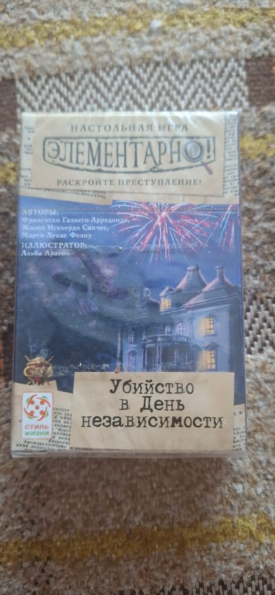 A vertically oriented, eye-level, slightly angled medium shot shows a board game box resting on a brown, textured fabric surface. The box is predominantly blue and depicts a dark, ominous house with lights on in the windows, set against a backdrop of fireworks. The title of the game, "Убийство в День Независимости" (Murder on Independence Day), is prominently displayed in a distressed font across the bottom half of the box, overlaying a tan-colored banner. Above the title, the game's name "ЭЛЕМЕНТАРНО!" (Elementary!) is written in a large, stylized font with a magnifying glass icon. Text indicating authors and illustrators is visible on the left side of the box. A small circular logo with abstract red and white figures is located in the lower-left corner of the box, next to the text "СТИЛЬ ЖИЗНИ" (Lifestyle). The fabric surface appears to be a rug or blanket with a repeating geometric pattern in shades of brown and yellow. There is no indication of the city of Tsimlyansk, Russia, in the image.