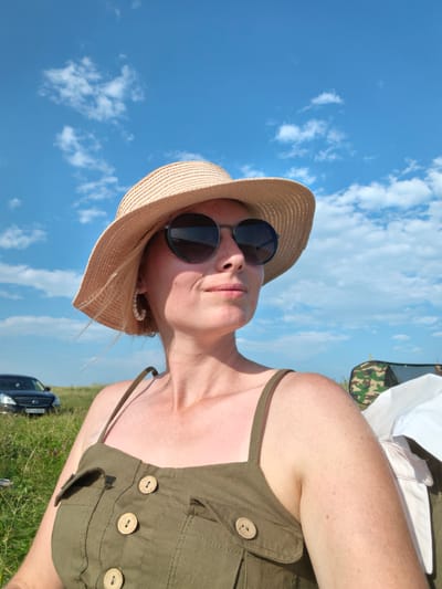 In Кузьминовка, Russia, under a bright blue sky with scattered white clouds, a woman with blonde hair, wearing a straw hat and sunglasses, is pictured from the chest up. She is also wearing a green sundress with visible wooden buttons and pearl hoop earrings. Her skin appears sun-kissed, and she is looking to her right. In the background, a dark car is parked in a grassy field, and a camouflage tent is visible to the right. The scene suggests an outdoor activity, possibly a picnic or a break during travel.