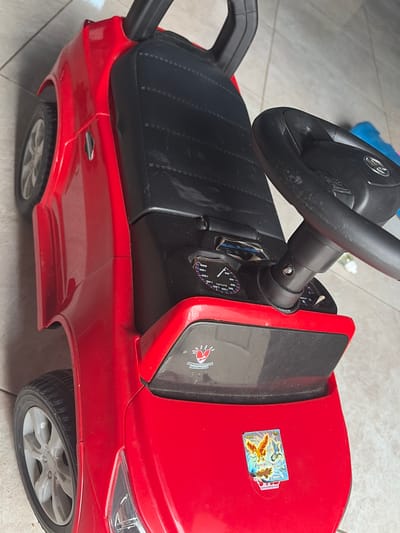 This is a close-up, overhead shot of a red toy car. The car is a ride-on toy with a steering wheel, a black seat, and a dashboard. The dashboard has a speedometer, with markings up to 200 km/h. The car also has toy wheels and a handle behind the seat, suggesting it's designed for a child to push or be pushed.

On the hood of the car, there are two stickers. One sticker appears to be from "PMB Toys" and features Indonesian text that translates to "Proudly made in Indonesia." The other sticker is a colorful holographic image of Pokémon characters, specifically Moltres and Zapdos.

The scene appears to be indoors or in a covered outdoor area, with a tiled floor visible in the background. The lighting suggests daytime. No people are visible in the image.