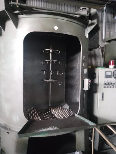 The image shows a large, industrial-style oven or drying chamber with a tiered rack inside, designed to hold items for processing. The rack has multiple levels, each with several hooks or prongs. A perforated metal sheet is positioned at the bottom of the chamber, possibly to catch debris or facilitate airflow. To the right of the oven is a control panel with various buttons, switches, and a digital display, indicating it's part of an automated industrial process. A red warning light is visible on top of the control panel. The overall environment appears to be a factory or workshop setting, with corrugated metal walls visible in the background. The image does not contain any people or provide specific location details for Sukaresmi, Indonesia.