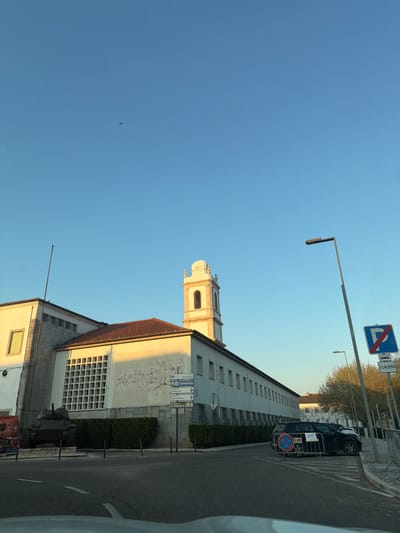 The image displays an outdoor street scene in Santarém, Portugal, situated near the Biblioteca Municipal Braamcamp Freire and the Convento de São Francisco. The setting is daytime under a clear blue sky, with warm, low-angle sunlight illuminating the scene from the right.
The central feature is a large, multi-story building with a white facade and a terracotta-tiled roof. A prominent square bell tower with arched openings and a decorative top rises from the building. To the left of the bell tower, a tall flagpole extends upwards. A section of the building's facade features a large, rectangular relief sculpture depicting multiple human figures in a dynamic, possibly historical, composition. Below this relief, there is a grid-patterned architectural element.
In front of the building, at street level, a dark green M4A3 Sherman tank, identified as the Monumento aos Combatentes do Ultramar, is positioned on a low stone plinth, partially obscured by neatly trimmed green hedges. To the immediate left of the tank, a portion of a red vehicle is visible.
The foreground consists of an asphalt road with white painted markings, including directional arrows and dashed lines. The road is bordered by a concrete curb lined with multiple vertical black bollards. On the right side of the road, metal crowd control barriers enclose a section of a paved area where a dark-colored SUV or station wagon is parked. A sign affixed to the barriers near the vehicle reads "C.M.S.". A white car is partially visible behind the dark SUV.
Street furniture includes a tall, modern street lamp pole on the right. Two informational street signs are also present: a blue square sign with a red diagonal bar indicating "No Parking" or "No Stopping", featuring the text "TURISMO" and a small bus/truck icon; and a white rectangular directional sign with multiple arrows pointing to "biblioteca municipal", "bomberos", "justiça II", "tribunal da concorrência", and "convento de S. Francisco II". Sparse, leafless trees are visible in the background on the right. A single, small, dark bird is visible in flight in the upper-mid section of the clear blue sky. No people are visible in the scene.