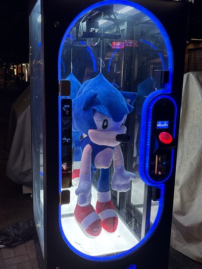 A slightly angled, medium shot depicts a Sonic the Hedgehog plush toy inside a prize vending machine, possibly a claw machine, with a blue neon-lit frame. The machine features a control panel with a red button and a small digital display that reads "Insert Coin" in a small black panel. A label with instructions on how to operate the machine is visible at the bottom, explaining the function of the forward button to move the scissors closer to the prize and the automatic cutting of the wire. The machine's exterior is black, and the background is dark. The location appears to be outdoors, potentially in Vrnjacka Banja Municipality, Serbia, considering the request in the prompt. No people are visible.