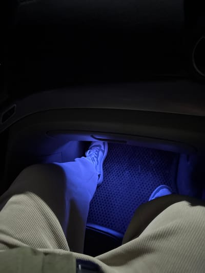 Inside a vehicle, a leg clad in light-colored pants and a sneaker rests on a floor mat illuminated by a vibrant blue light. The car's interior, mainly the door and dashboard area, is visible. The lighting creates a strong contrast, with the leg and floor bathed in bright blue, while the rest of the car's interior fades into near darkness. The focus is on the lower half of the body and the immediate surrounding area.