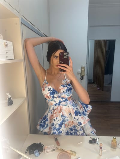A young woman with dark hair is taking a selfie in a well-lit room, possibly a dressing room or bedroom. She is wearing a floral print dress with blue and pink flowers. Her left arm is raised and resting on her head, and she is leaning against a white wall. In the foreground, on a white surface that appears to be a vanity or desk, there are various items, including a black digital camera, makeup, keys, and coins. In the background, a large mirror reflects part of the room, showing wooden paneling and a door. The overall environment appears to be indoors, with clean, modern furnishings. The exact location is not specified as Agios Dimitrios, Greece, or any other city.