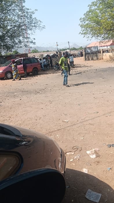 The image depicts an outdoor scene in Langtang, Nigeria, under a clear daylight sky. The immediate foreground shows the dark-colored hood and side mirror of a vehicle, indicating the perspective. The ground is an unpaved, light brown dirt surface, littered with scattered debris including paper and plastic.
In the midground, a red minivan is stopped on the left. Two individuals are positioned near it; one person, possibly a female, wears a multi-colored patterned dress and stands by the driver's side door, while another figure is partially obscured near the vehicle's rear. To the right of the minivan, a group of approximately ten individuals, predominantly male, are gathered, with several seated on or standing next to at least five motorcycles. A male individual in a lime-green t-shirt and blue jeans stands prominently in the center of the dirt area, casting a distinct shadow to his right.
The background features a building with a reddish-brown roof and a dark, possibly fenced, perimeter on the right. On the left, a large tree with green foliage stands, partially obscuring a red and white lattice telecommunications tower. Distant hills or mountains are visible under the pale blue sky.