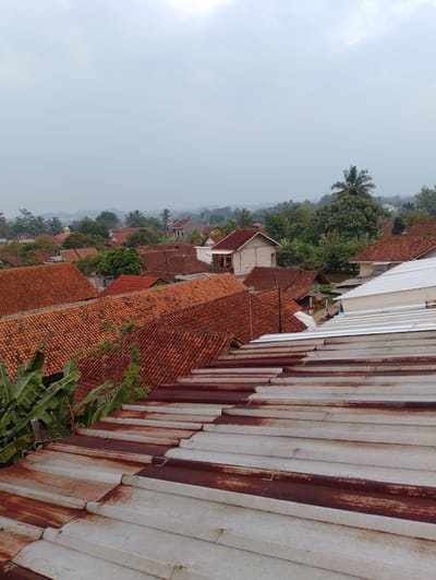 The image captures a view of rooftops in Sukamanah, Indonesia. In the foreground, weathered corrugated metal roofing, showing signs of rust, extends into the frame. Beyond this, a dense expanse of reddish-orange clay tile roofs covers numerous houses. Several banana trees with large green leaves are visible in the mid-ground to the left, partially obscuring some of the tiled roofs. Further back, the landscape is dotted with more homes, interspersed with lush green trees and vegetation. A yellow dome, possibly belonging to a mosque, is visible in the distance amongst the trees. The sky is overcast with a pale, diffused light, suggesting a cloudy day. The overall scene depicts a residential area with traditional architecture and abundant tropical foliage.