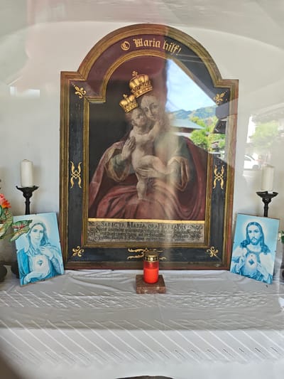 The image shows an indoor shrine or niche, likely located in Kitzbühel, Austria, given the context provided. The central focus is a framed painting of the Madonna and Child, both wearing crowns. Above the painting, in gold lettering on a dark background, is the inscription "Maria hilf!". Below the painting, there is a gold-colored decorative border framing a text that is partially obscured by glare and reflection but appears to be in Latin.
To the left of the central painting, a framed image of the Virgin Mary, depicted in blue and white, is propped up. To the right, a similar framed image of Jesus Christ is also propped up, with his hands held over his chest.
In front of the central painting, a red votive candle in a glass holder sits on a small marble slab. Two white pillar candles in dark, ornate candle holders are positioned on either side of the shrine, flanking the framed images. The shrine is covered by a white tablecloth with a decorative, patterned overlay. The background wall is white, and the overall lighting suggests an indoor environment with some natural light coming in, causing reflections on the glass of the central painting. The presence of religious imagery and votive candles indicates this is a devotional space.
