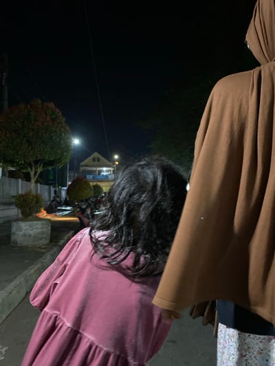 This is a nighttime photograph taken in the city of Tanggul, Indonesia, on a street. The image shows the back of two individuals, likely a child in the foreground and an adult behind them. The child is wearing a pink dress with ruffled detailing. The adult is wearing a brown headscarf and a brown garment. In the background, there are trees, a building with lights on, and a streetlamp. The scene appears to be outdoors, possibly in a residential or commercial area, with evidence of human activity such as people sitting on the ground and motorcycles parked. The overall atmosphere is dark and illuminated by artificial lighting.