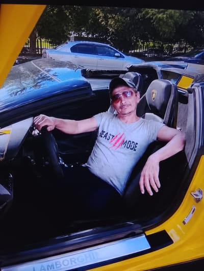 The media file shows a man sitting in the driver's seat of a yellow convertible car, which appears to be a Lamborghini. The man is wearing a gray t-shirt with the text "BEAST MODE" printed on it in black and red. He also has on sunglasses and a baseball cap. The car's interior is black with yellow accents, and the Lamborghini logo is visible on the headrest of the seat. In the background, other cars and trees are visible, suggesting an outdoor setting. The lighting suggests it is daytime, and the presence of trees indicates a natural environment. The man appears relaxed, with a slight smile. The text "LAMBORGHINI" is visible on the door sill of the car. The overall impression is of a man enjoying a luxury vehicle.