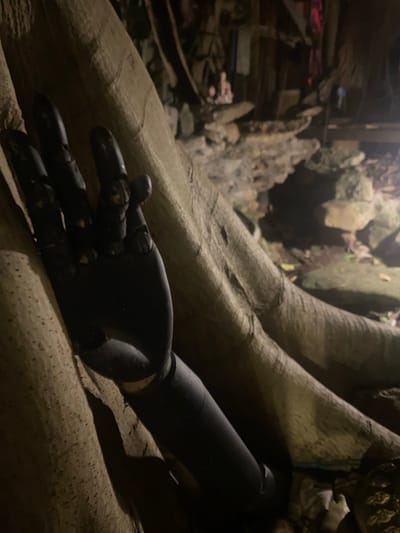 A black, articulated prosthetic arm lies on the textured surface of a large tree root in Baan Don Sai, Thailand. The hand of the prosthetic is positioned with slightly bent fingers. The environment is dimly lit, casting shadows across the scene. The background includes rocky terrain and foliage.