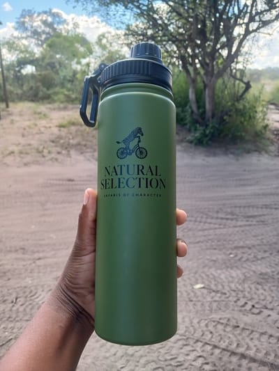 A person is holding a green water bottle with a black lid and a logo that reads "NATURAL SELECTION SAFARIS OF CHARACTER." The background shows a dirt road with tire tracks and vegetation on either side, including trees and grass. The location is unspecified; the city of Nwanaeanoka, Botswana is not verifiable within the visual context.