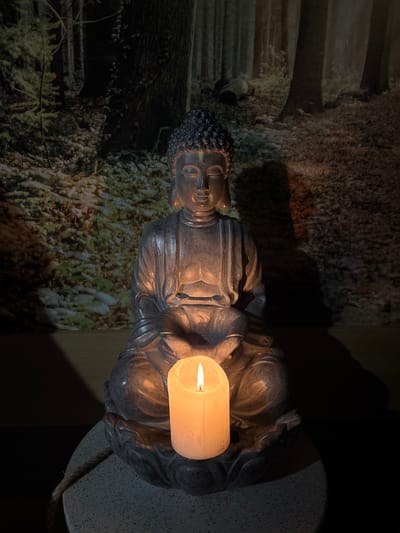 The image displays an indoor scene in Баня, Bulgaria. A dark grey, seated Buddha statue occupies the central foreground, rendered in a meditative posture with closed eyes and hands joined in Dhyana Mudra, resting on its lap. A thick, off-white pillar candle, actively burning with an orange flame, is positioned directly within the Buddha's cupped hands, serving as the primary light source. The candle's light illuminates the lower portion of the statue and casts a warm glow upwards onto its torso and face. The statue rests on a circular, light-colored, speckled base, from which a braided rope or cord descends on the left side. The background consists of a wall mural or wallpaper depicting a dense forest with numerous dark tree trunks and a forest floor covered in green vegetation, possibly frosted or lightly snow-dusted, under dim lighting conditions. No individuals are present in the scene.