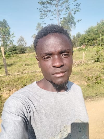 A man with dark skin, a short beard, and short, dark hair wears a gray T-shirt. He is outside in a grassy area with trees and a light blue sky. The photo appears to be a selfie taken at an angle, causing the man to appear slanted. The lighting is bright, suggesting it is daytime. A shadow is cast on the side of his shirt.