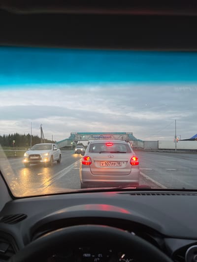 The image captures a view from inside a car, looking out at a traffic jam on a cloudy day, likely in the late afternoon or early evening, given the dim light. The road appears wet, suggesting recent rain. Several cars are visible, including a silver car in the foreground with its brake lights illuminated, and a white car further back with its headlights on. A pedestrian bridge spans the road in the distance. The scene is set on a multi-lane road, possibly a highway or major thoroughfare, with trees lining the sides. The sky is overcast with a mixture of grey and white clouds. The interior of the car is partially visible, including a dark-colored dashboard and steering wheel.

Visible text includes a license plate on the silver car which reads "У 107 РР 18 RUS". There is also some unreadable text on a screen or display within the car's interior on the right side of the image.