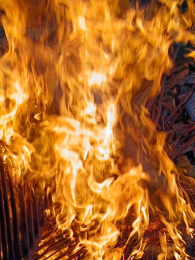 The image is a close-up of a fire engulfing a grill, with flames dominating the frame in hues of orange and yellow. Underlying the intense heat, faint outlines of thin, dark, vertical bars of the grill are visible on the left side. In the background, there are indistinct shapes of debris, possibly dry leaves and twigs, adding to the impression of an outdoor fire. The setting is a city in Indonesia, specifically Meuliek. The image provides no information about people, specific actions, or interactions beyond the presence of the fire and the grill.