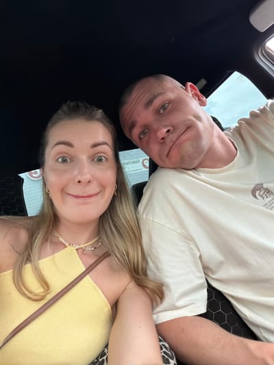 Two individuals, a woman and a man, are captured in a selfie taken from within a vehicle. The woman, positioned on the left, has long blonde hair and is looking directly at the camera with wide, surprised eyes and an open-mouthed smile. She is wearing a yellow top and a beaded necklace. The man, on the right, has a shaved head and is leaning his head against the woman's shoulder, looking towards the camera with a slight smirk. He is wearing a light-colored t-shirt. The background suggests the interior of a car, with a dark ceiling and a window showing an overcast sky. The location is identified as Gubarevo, Russia.