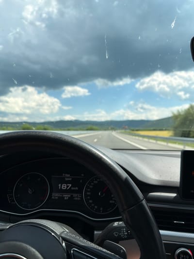 The image shows the view from inside a car driving on a highway in Peshterna, Bulgaria. The car's dashboard is visible, displaying a speed of 187 km/h. Outside, the road stretches into the distance, flanked by green hills under a sky with dramatic clouds. The windshield appears to have some minor streaks on it.