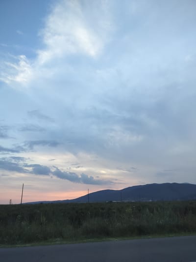The image depicts a serene rural landscape at dusk, with the sky painted in soft hues of blue, gray, and a hint of peach near the horizon, suggesting the last moments of daylight. Wispy clouds drift across the vast expanse, creating a sense of depth and tranquility. In the foreground, a dark asphalt road runs along the bottom edge of the frame, hinting at a paved pathway. Beyond the road, a lush field of tall grass and wildflowers stretches towards a distant mountain range. The mountains are silhouetted against the fading sky, their dark forms creating a striking contrast. A few sparsely placed utility poles stand like sentinels in the field, adding a touch of human presence to the natural scenery. The overall mood of the image is peaceful and contemplative, evoking a sense of quiet solitude and the beauty of nature as day transitions into night. The location is identified as Manolovo, Bulgaria.