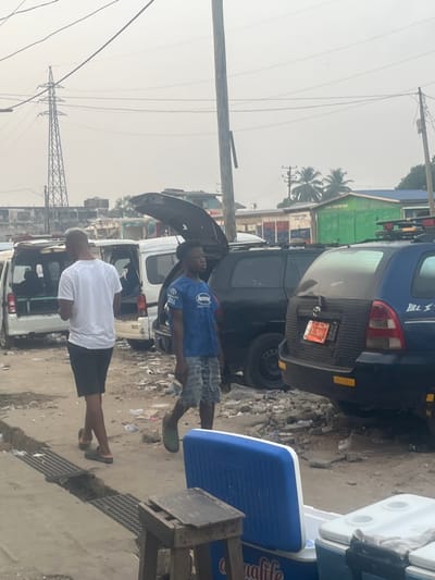 A street scene shows two men standing near vehicles with open hoods. One man wears a white shirt and black shorts, while the other wears a blue t-shirt and patterned shorts. Debris litters the ground. A blue SUV with a visible license plate is parked nearby. Power lines and a utility pole stretch across the sky, with palm trees in the background. A cooler rests on a makeshift stand in the foreground.